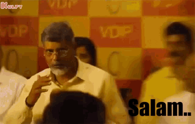 a man with glasses and a beard is standing in front of a crowd and saying salam .