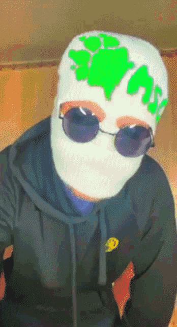 a person wearing a mask and sunglasses has a green sticker on their hat that says trash