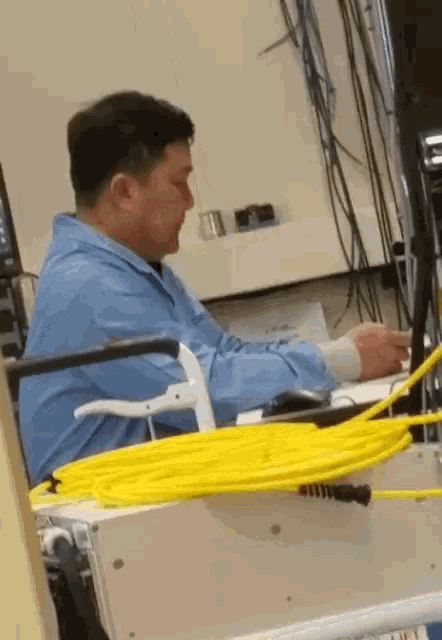 a man in a blue lab coat sits at a desk with a bunch of wires