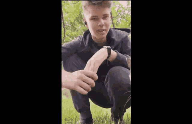 a young man is squatting down in the grass with a watch on his wrist .