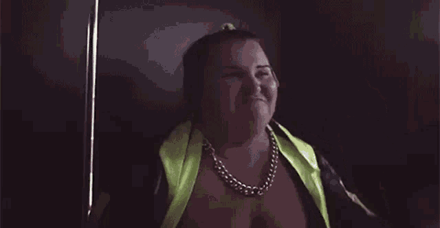 a woman is standing in a dark room wearing a yellow jacket and a gold chain .