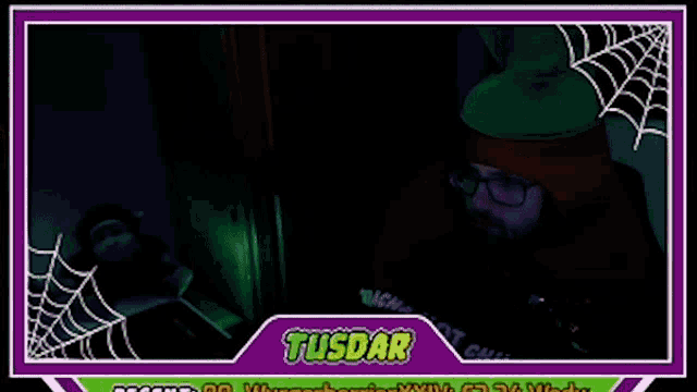 a man in a red hoodie and glasses is sitting in a dark room with a spider web in the background .