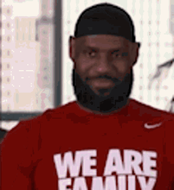 a man with a beard is wearing a red shirt that says `` we are family '' and a black hat .