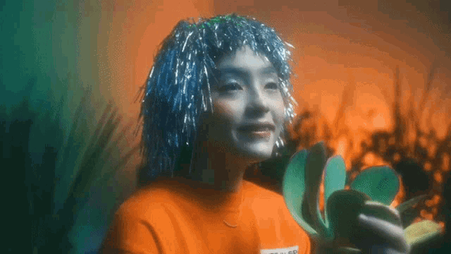 a woman wearing a blue wig and an orange shirt holds a cactus