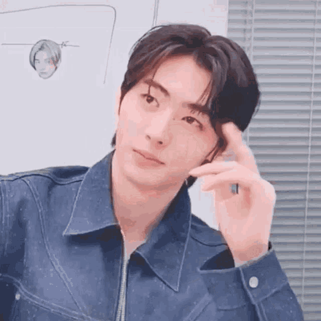 a young man in a denim jacket is looking at the camera while holding his hand to his forehead .