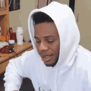 a man in a white hoodie is sitting in front of a desk .