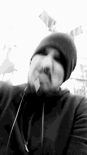a man with a beard wearing a beanie and headphones is smoking a cigarette in a black and white photo .