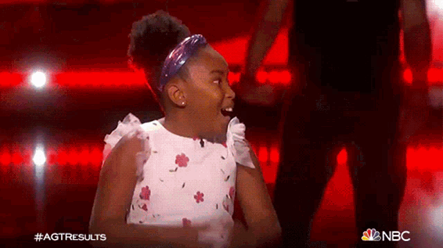 a little girl in a white dress with flowers on it is on a stage with a man in a black shirt behind her .