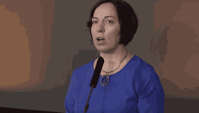 a woman in a blue shirt stands in front of a microphone with a pendant that says ' volvo ' on it