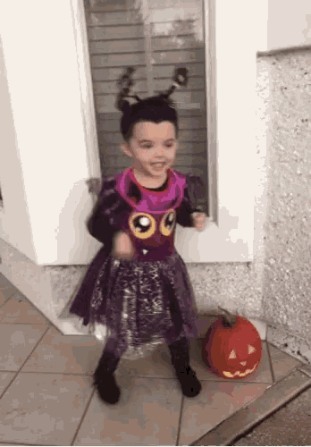 a little girl dressed in a purple costume stands next to a pumpkin