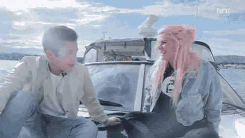 a woman with pink hair is sitting next to a man on a boat with the letters nrk visible in the background