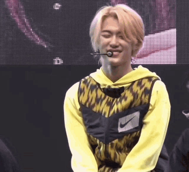 a young man wearing a yellow hoodie and a black vest with a microphone in his mouth is sitting on a stage .