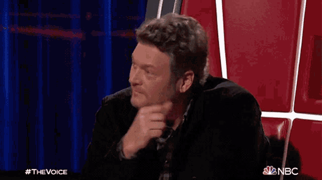 a man in a black jacket is sitting in a chair with his hand to his chin .