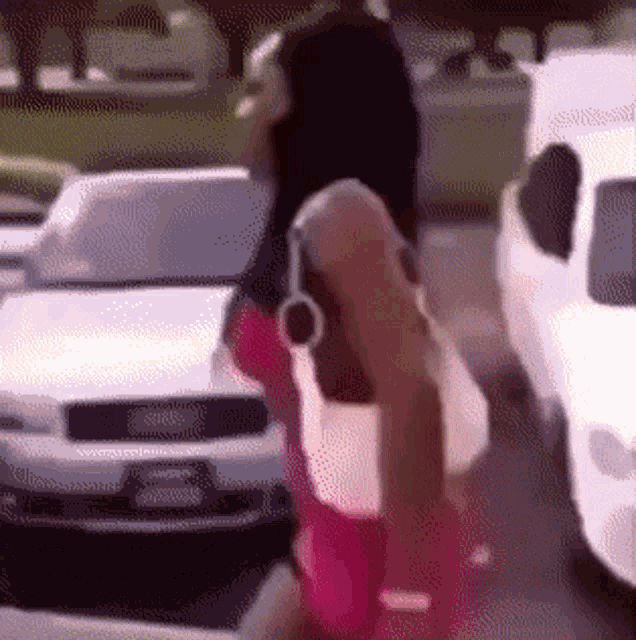 a woman in a pink dress is walking down a street in front of a white car