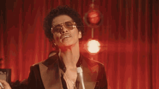 a man wearing sunglasses and a suit stands in front of a red curtain with the word this written on it