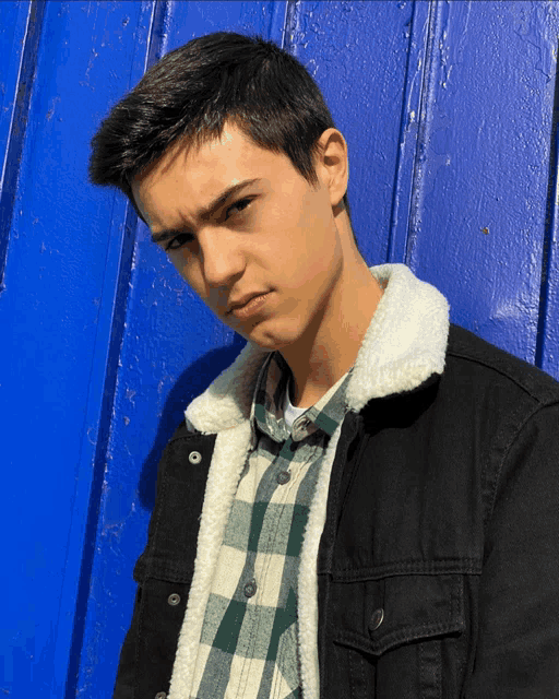 a young man wearing a black jacket and a plaid shirt is standing in front of a blue wall