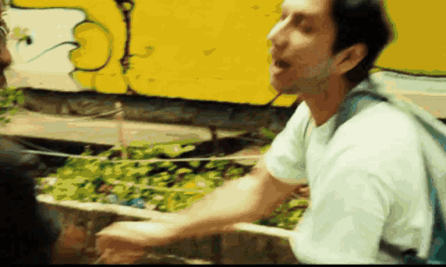 a man in a white shirt is talking to another man in front of a yellow wall with graffiti on it