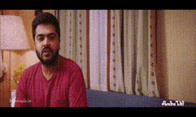 a man with a beard is sitting on a couch in front of a window in a room .