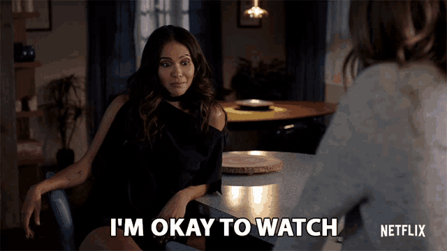 a woman sitting at a table with the words i 'm okay to watch on the bottom