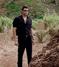 a man wearing sunglasses and a black shirt is standing on a dirt path in a field .