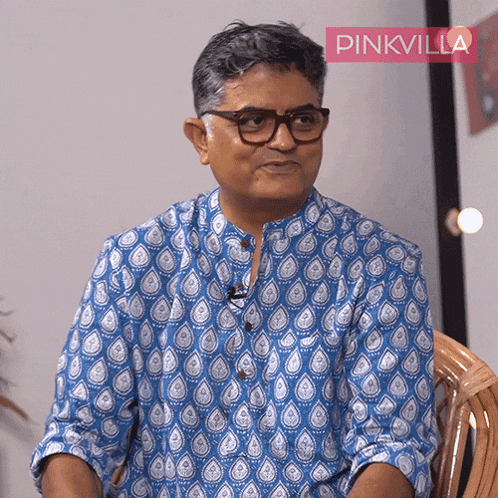 a man wearing glasses and a blue shirt is sitting in a chair with a pinkvilla sign behind him
