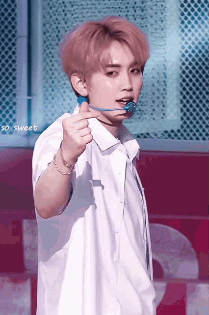 a young man with pink hair is wearing a white shirt and holding a blue object in his mouth