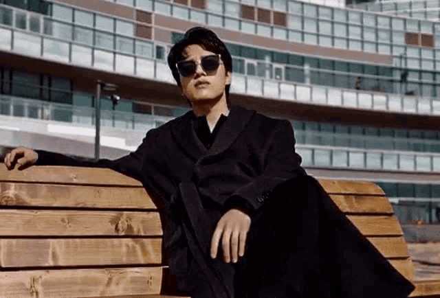 a man wearing sunglasses and a black coat is sitting on a bench in front of a building .