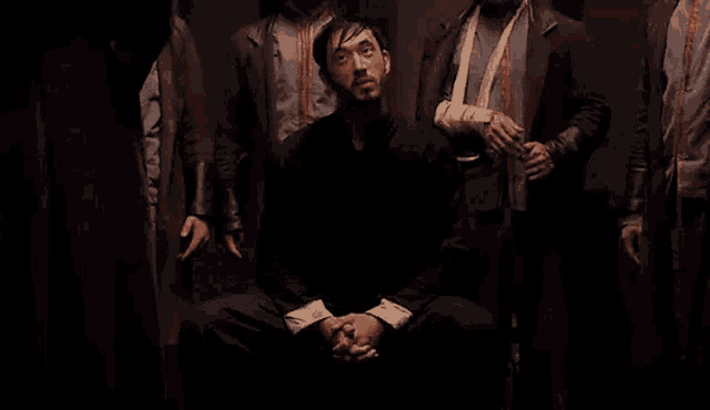 a group of men are standing around a man sitting in a chair in the dark .