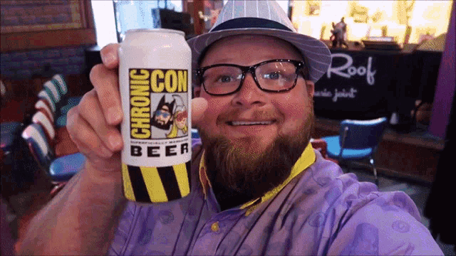 a man wearing glasses and a hat holds up a can of comic con beer