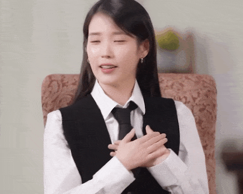 a woman in a vest and tie is sitting in a chair with her hands on her chest