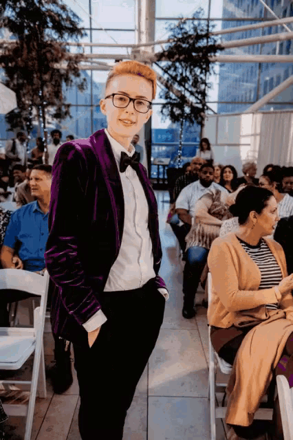 a woman in a purple jacket and bow tie stands in front of a crowd of people