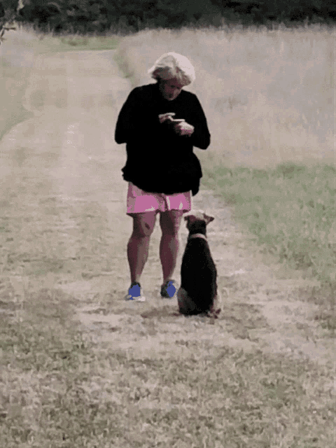 a woman in pink shorts stands next to a black dog on a dirt path