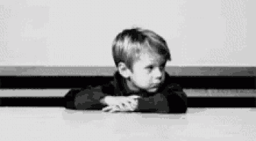 a black and white photo of a young boy sitting at a table with his hands on his chin .