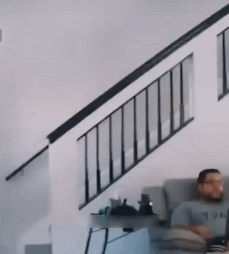 a man is sitting on a couch in front of a staircase while a cat is flying down the stairs .