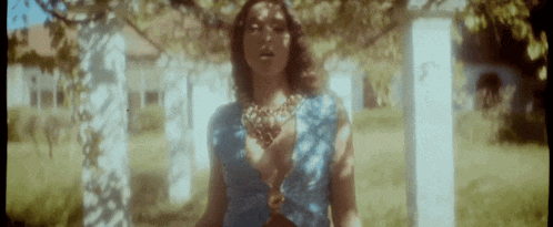 a woman in a blue dress is standing in a field with a necklace around her neck .