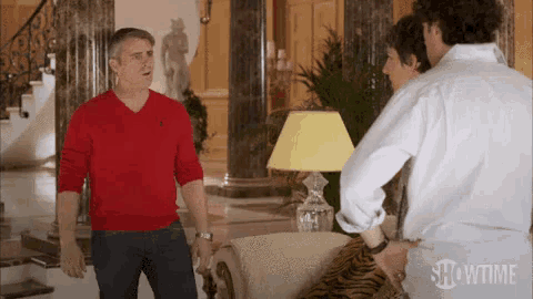 a man in a red sweater is standing in a living room talking to a woman and a man in a white shirt .