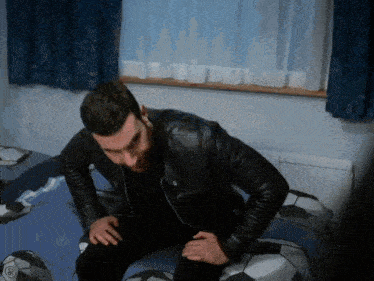 a man in a black leather jacket is sitting on a bed