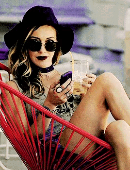 a woman wearing sunglasses and a hat is sitting in a red chair holding a drink and looking at her phone