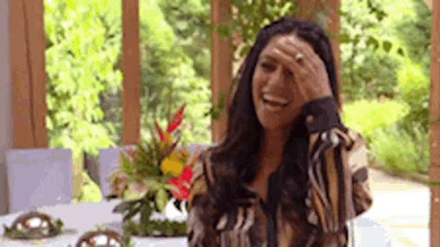 a woman is laughing while sitting at a table with flowers in the background .