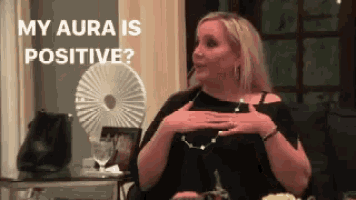 a woman is sitting at a table with her hands on her chest and the words `` my aura is positive '' behind her .