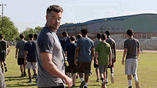 a man in a grey shirt is standing in front of a group of people walking in a field .