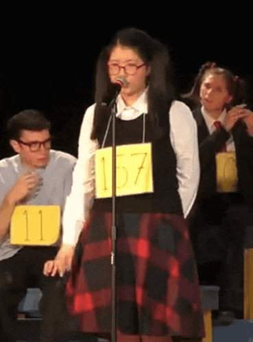 a girl in a plaid skirt is singing into a microphone while wearing a yellow tag that says 157