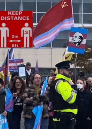 a man in a yellow jacket stands in front of a crowd of people holding flags and a sign that says keep distance