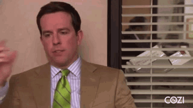 a man in a suit and green tie is sitting in front of a window with blinds .