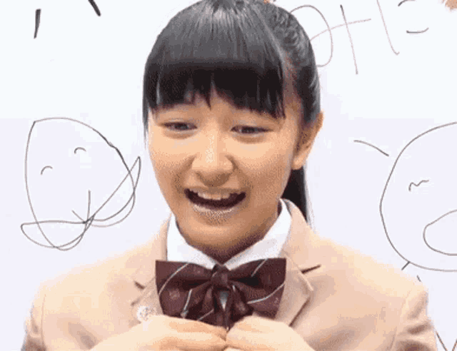 a girl in a school uniform is smiling in front of a whiteboard with drawings of faces on it .