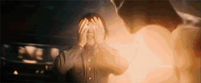 a man is covering his eyes in front of a fire while a man is standing in the background .