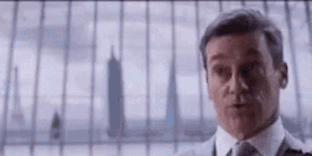 a man in a suit and tie is standing in front of a window with a skyscraper in the background .