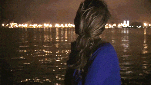 a woman in a blue sweater stands in front of a body of water at night