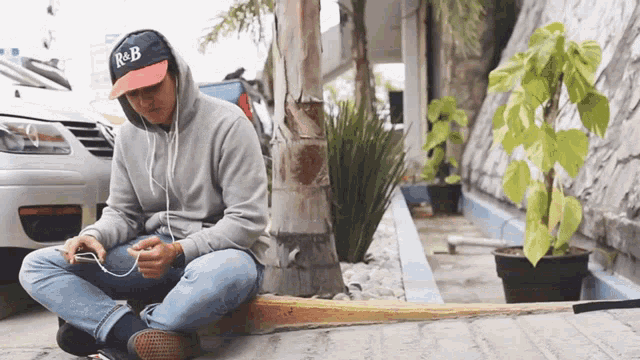 a man wearing a r & b hat sits on the sidewalk listening to music