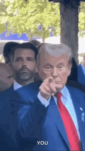 a man in a suit and tie is pointing at the camera while another man looks on .